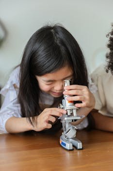 Cute little Asian girl with long dark hair making experiment with microscope while sitting at wooden table in classroom